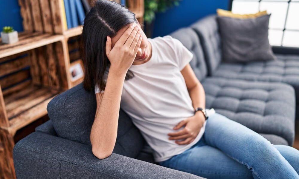 Mulher sentada em sofá com a mão na cabeça e outra mão na barriga.