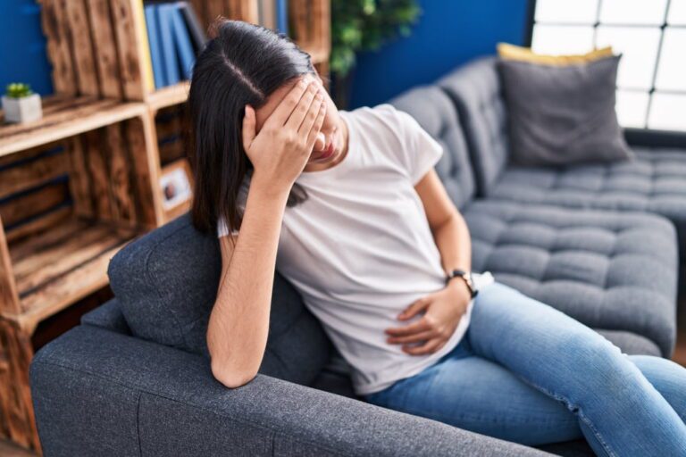 Mulher sentada em sofá com a mão na cabeça e outra mão na barriga.