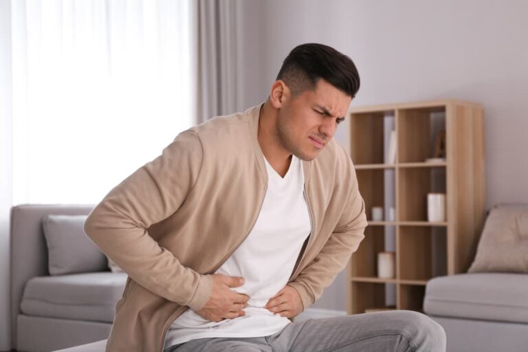 Homem sentado em um sofá segurando a área abdominal com expressão de dor, possivelmente indicando sintomas de hérnia.